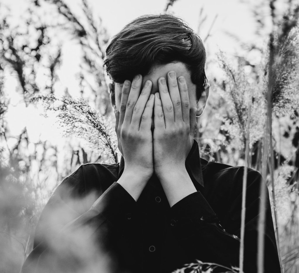 A black-and-white photo of a young man covering his face with his hands in a field, symbolizing the inner struggle and courage involved in embracing negative emotions.
