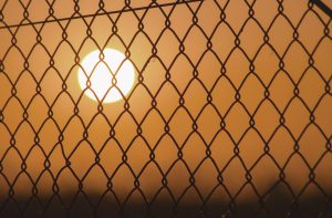 A glowing sun viewed through a rigid chain-link fence, illustrating why therapy isn't working when it tries to constrain human nature within artificial structures.