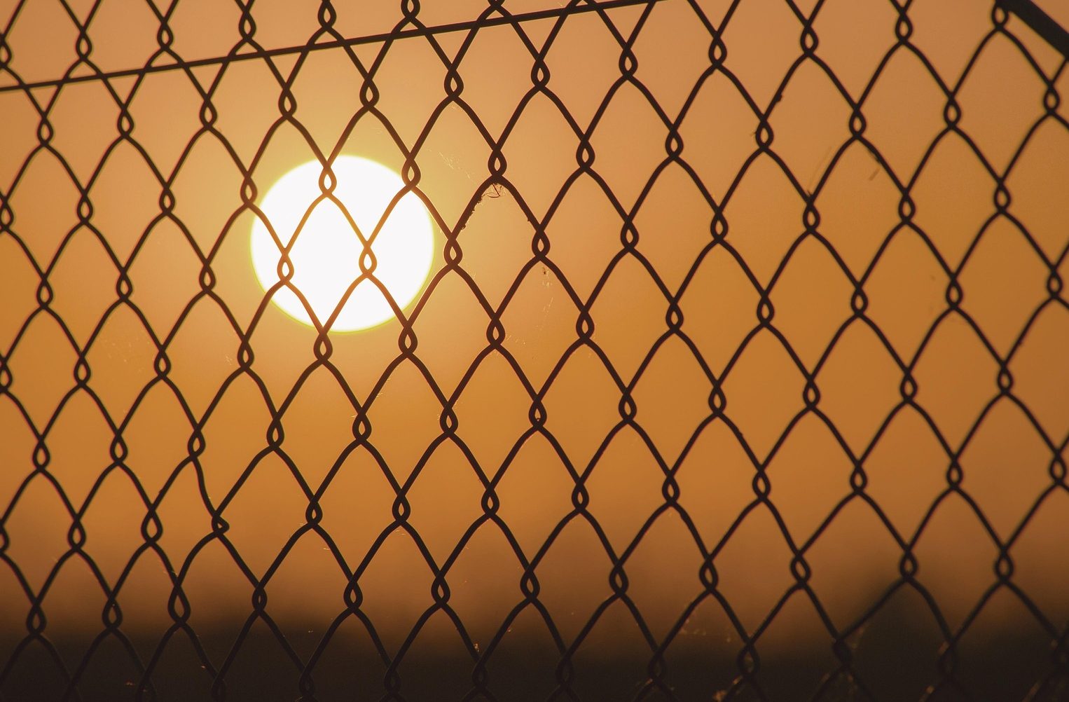 A glowing sun viewed through a rigid chain-link fence, illustrating why therapy isn't working when it tries to constrain human nature within artificial structures.
